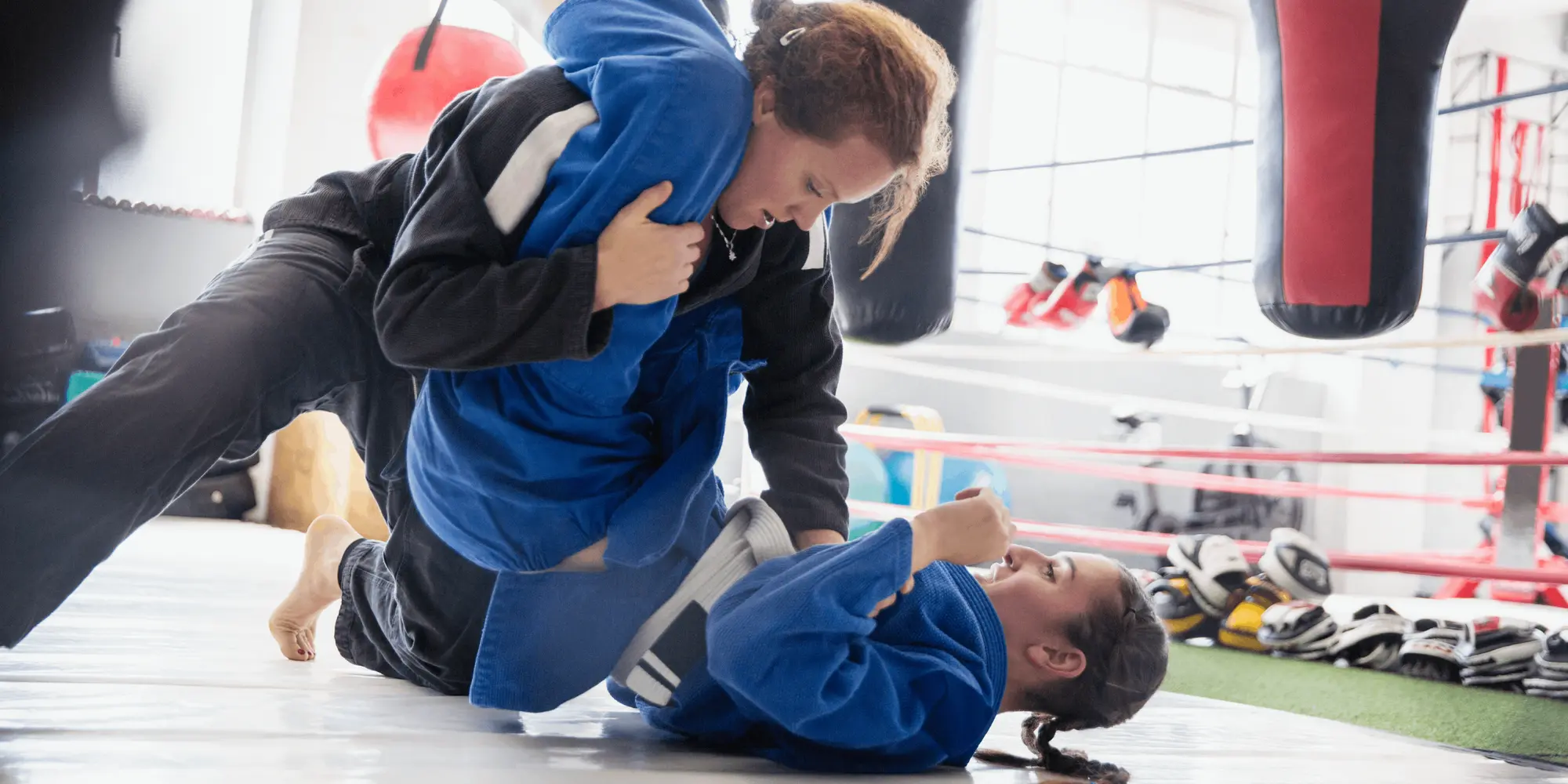 Två kvinnor tränar brasiliansk jiu-jitsu på en matta i en träningslokal. Den ena kvinnan ligger på rygg i blå dräkt och håller fast den andra kvinnan, som är klädd i svart dräkt, i ett grepp. I bakgrunden syns boxningssäckar och annan utrustning.