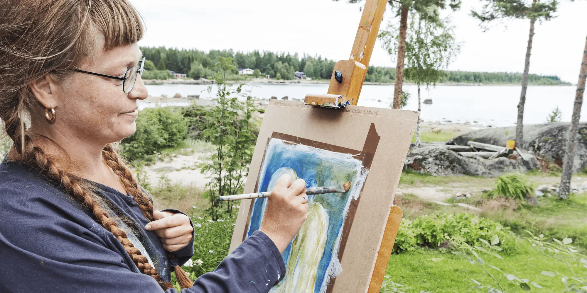 Kvinna målar vid staffli utomhus med sjö och skog i bakgrunden