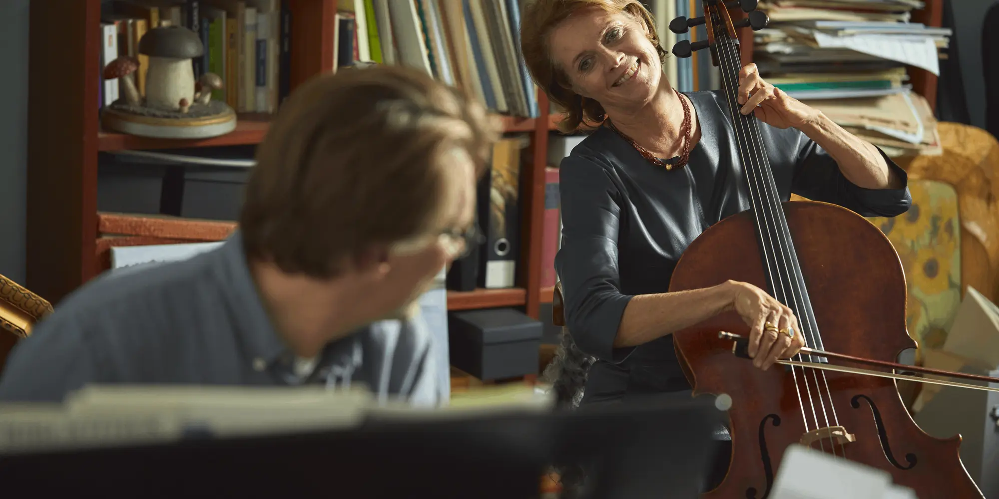 : En kvinna spelar cello med ett leende i ett rum fullt av böcker och papper. Hon bär en mörk topp och ett rött halsband.
