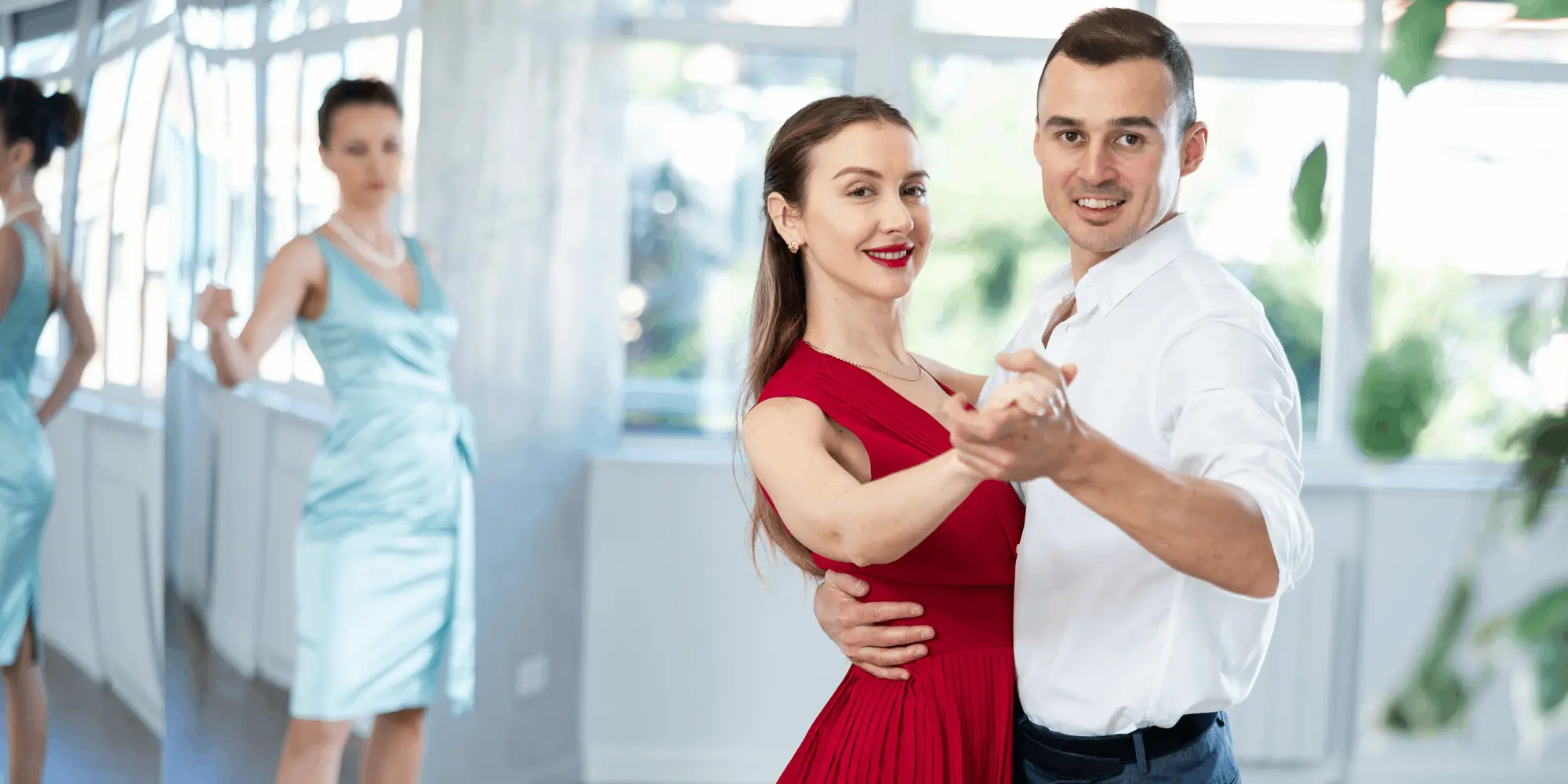 Ett par dansar i förgrunden – kvinnan i röd klänning och mannen i vit skjorta – medan en annan kvinna i ljusblå klänning står i bakgrunden och tittar på