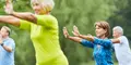 En grupp äldre personer tränar tai chi eller liknande rörelseövningar utomhus i en park. Fokus ligger på en kvinna i blå mönstrad tröja som står med armarna sträckta framåt. I bakgrunden syns fler deltagare i färgglada träningskläder.