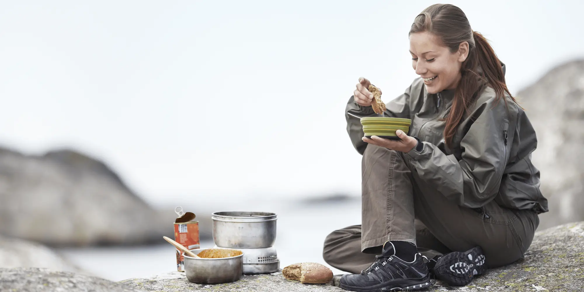 Leende kvinna i friluftskläder sitter på ett klipphäll vid havet och äter varm mat, omgiven av stormkök
