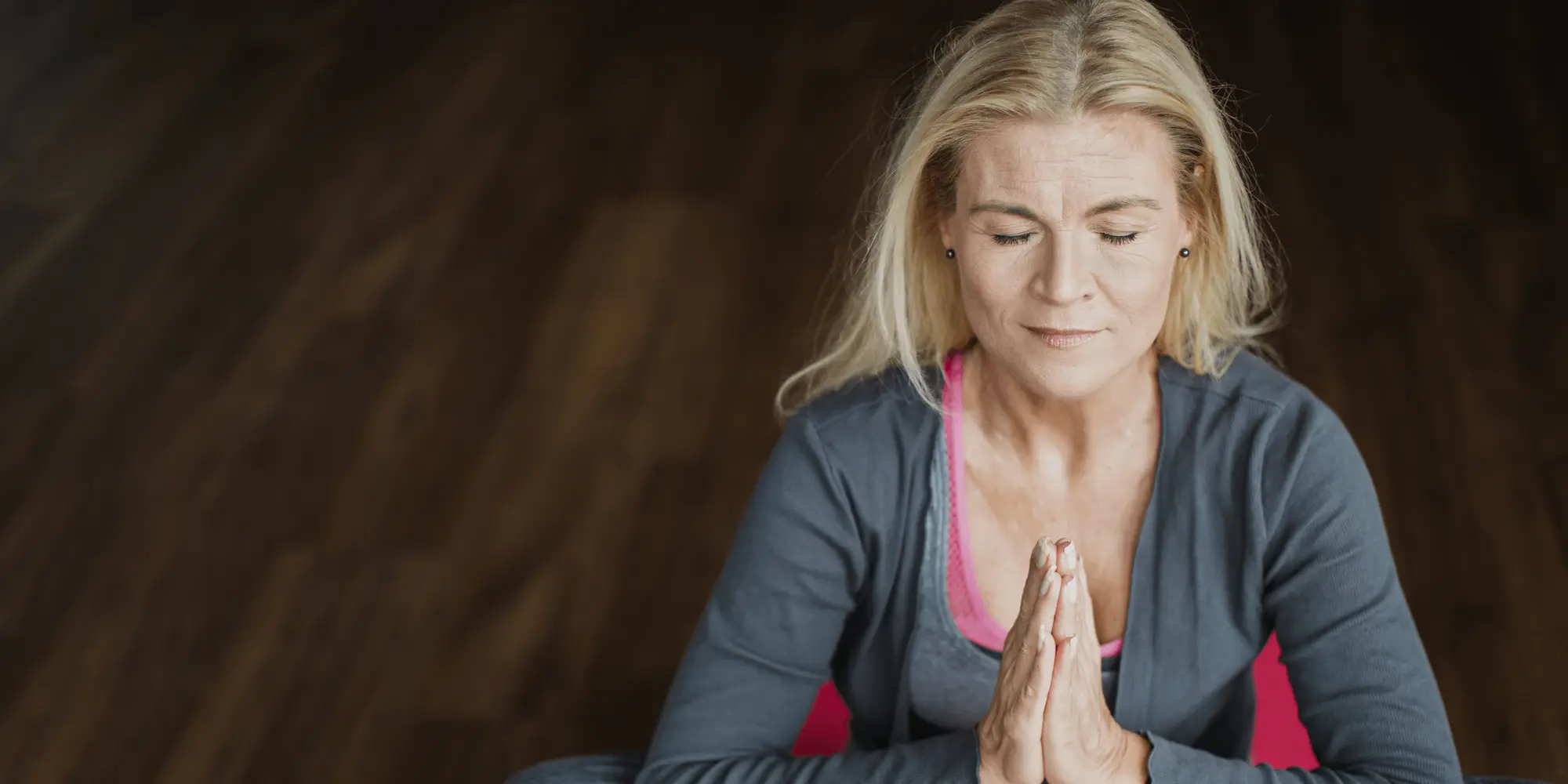 : Kvinna med långt blont hår sitter i skräddarställning på ett yogapass med ögonen slutna och händerna i böneposition framför bröstet.