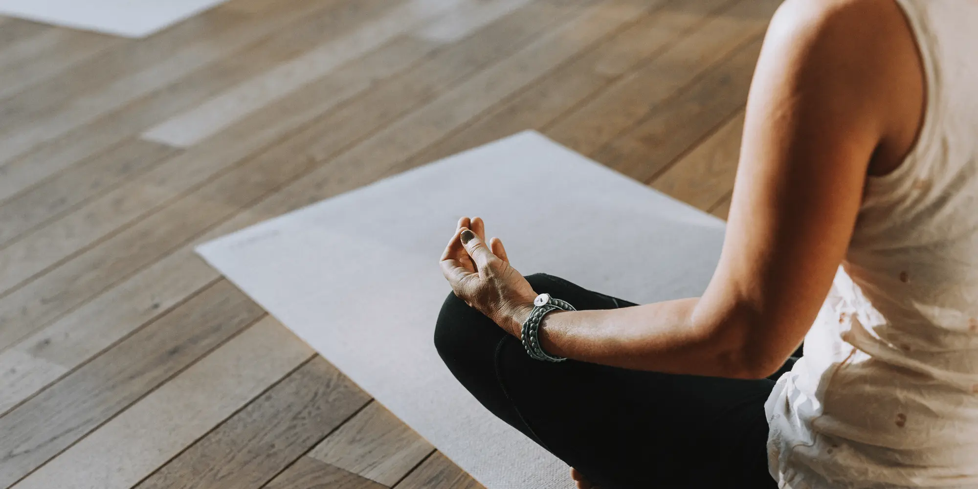 En person sitter i meditationsställning på en yogamatta i ett ljust rum med trägolv.
