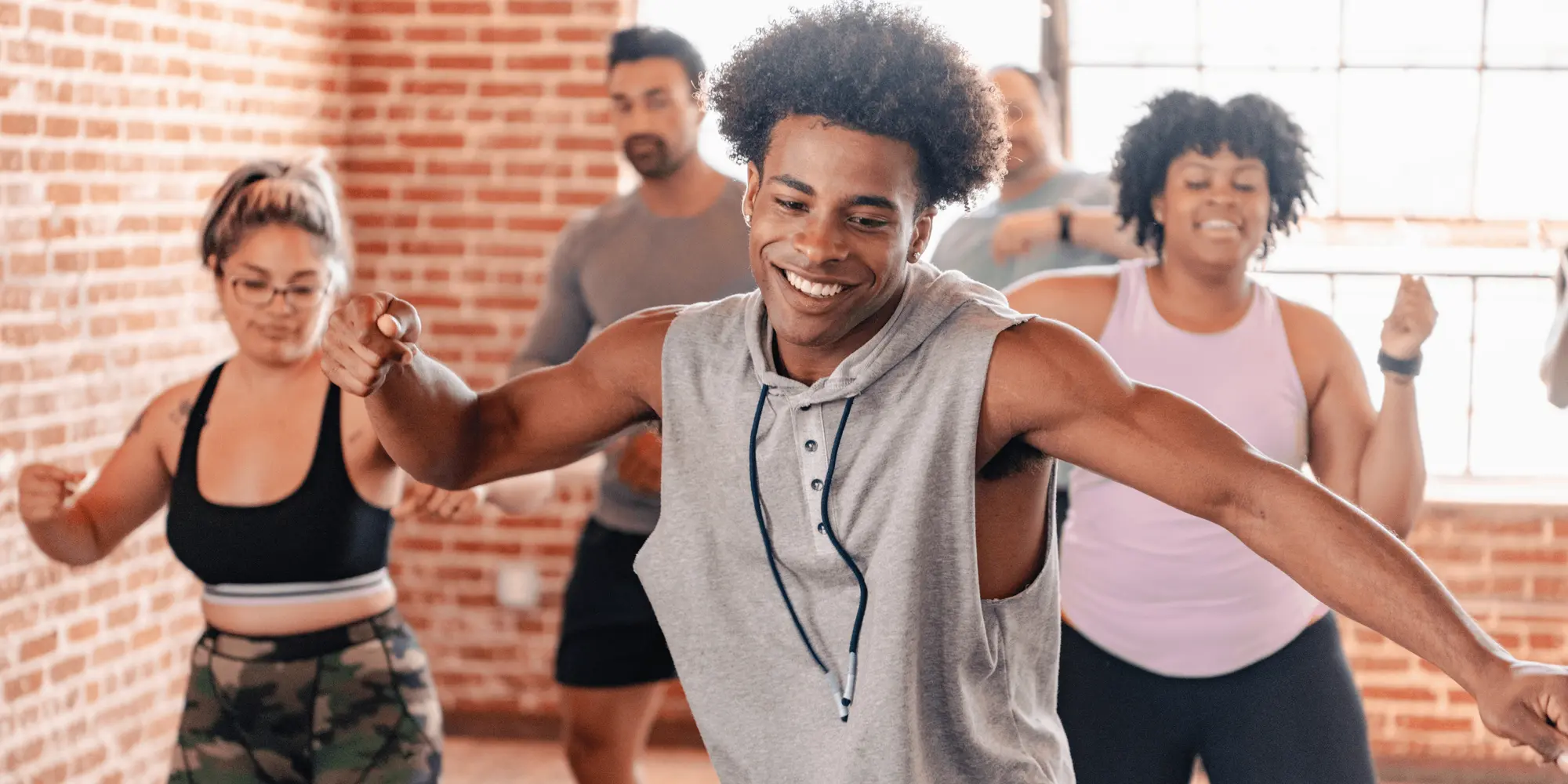 En grupp människor dansar energiskt tillsammans i en ljus dansstudio med tegelväggar. I förgrunden syns en leende man i grå ärmlös hoodie, och bakom honom dansar fyra andra deltagare med fokus och glädje