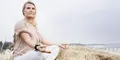 Kvinna sitter i meditationsställning i ett strandlandskap med högt gräs.
