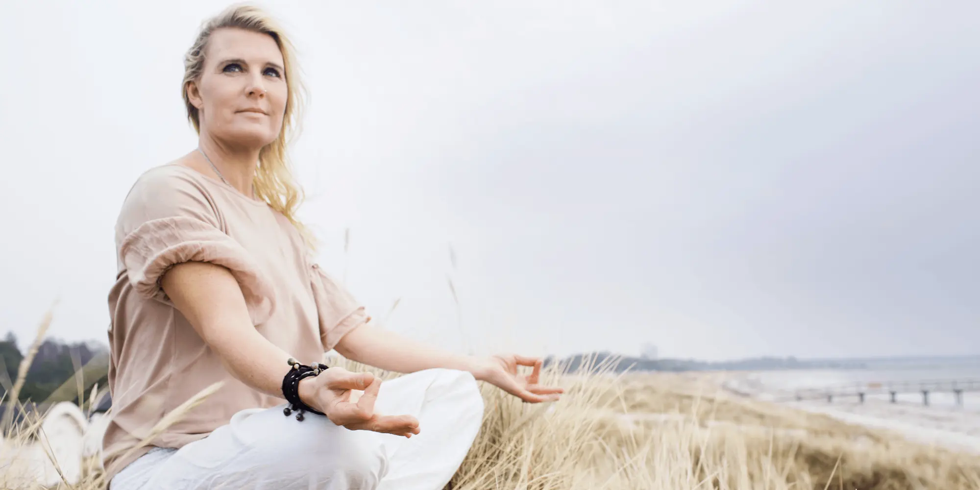 Kvinna sitter i meditationsställning i ett strandlandskap med högt gräs.