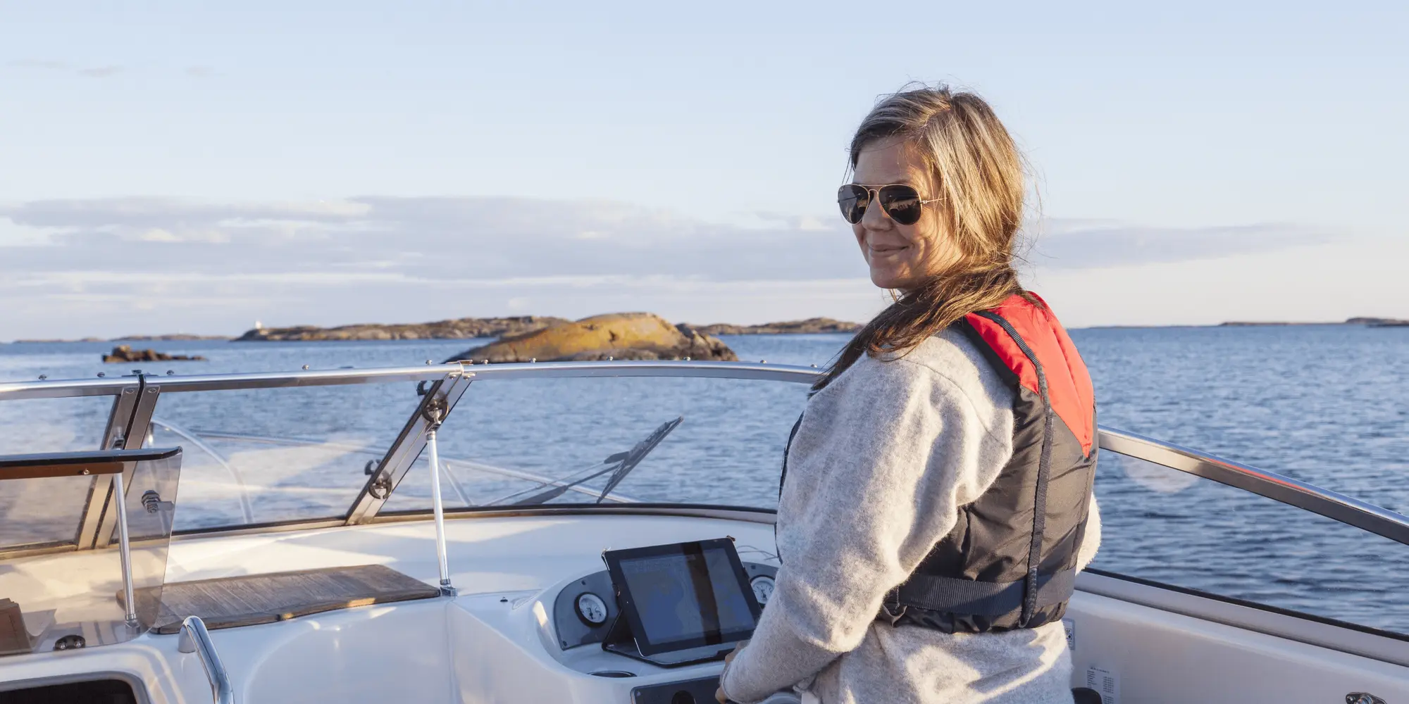 En kvinna med solglasögon och flytväst står vid rodret på en motorbåt, med skärgård och klippor i bakgrunden under en klar himmel