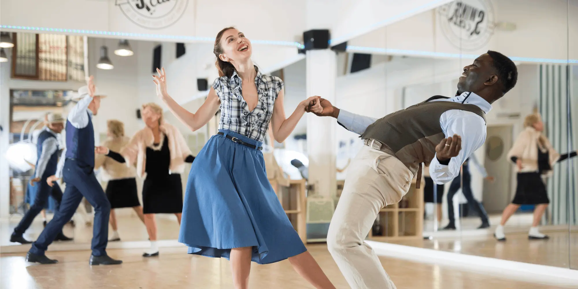 Par dansar energiskt lindy hop eller charleston i en spegelsal. Kvinnan bär blå kjol och rutig blus, mannen beige byxor och väst