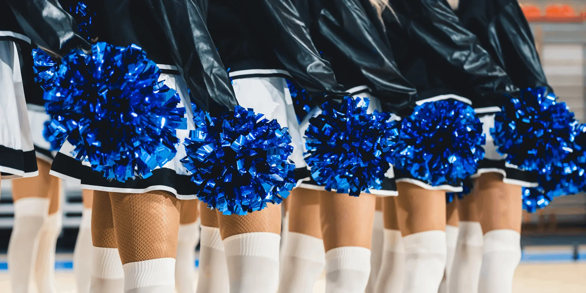 Cheerleaders står på rad i korta kjolar, vita knästrumpor och håller blå glittriga pompoms, redo för uppträdande inomhus i en sporthal