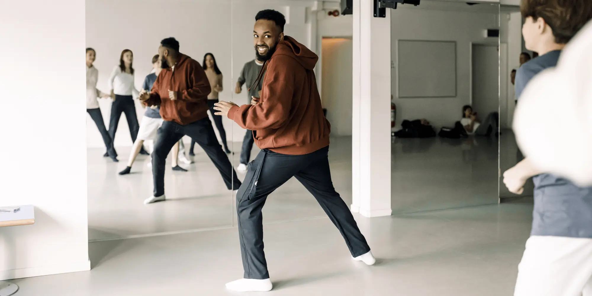 En danslärare i röd hoodie leder en grupp ungdomar i en ljus dansstudio med spegelväggar