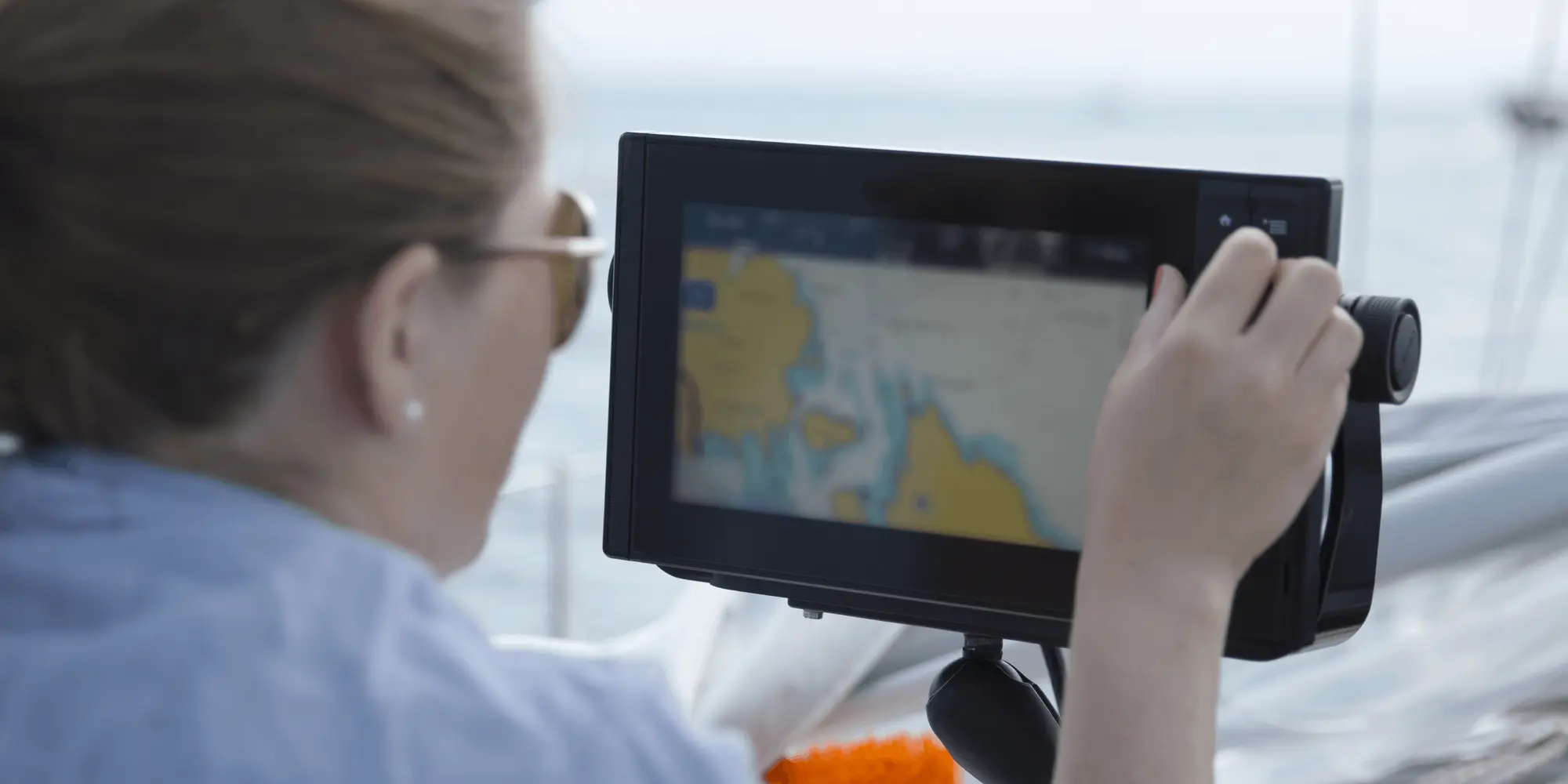 En kvinna med solglasögon och uppsatt hår navigerar en segelbåt genom att använda en digital sjökortsplotter.