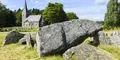 Stora stenblock i ett fornlämningsområde i förgrunden, med en gammal kyrka i sten och en kyrkogård i bakgrunden, omgiven av grönska och blå himmel