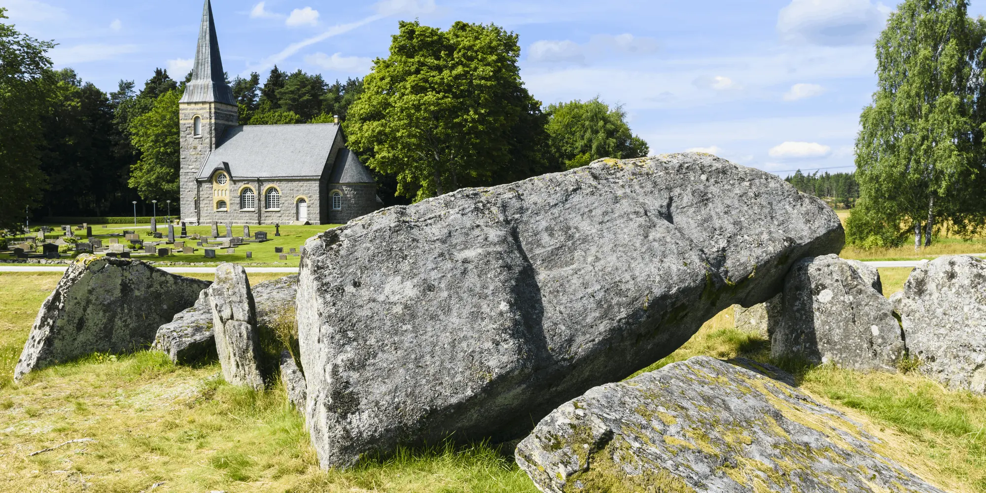 Stora stenblock i ett fornlämningsområde i förgrunden, med en gammal kyrka i sten och en kyrkogård i bakgrunden, omgiven av grönska och blå himmel