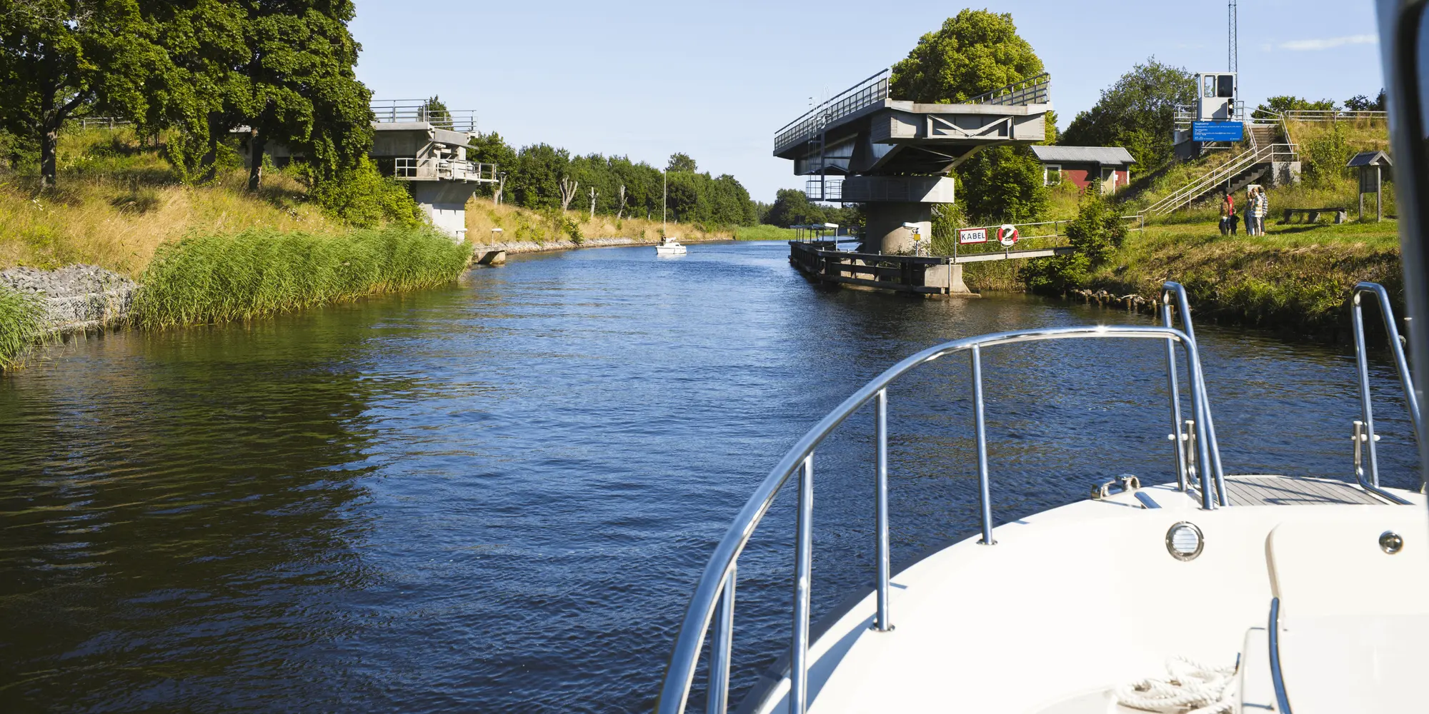 Utsikt från en båt som närmar sig en öppnad klaffbro på en smal kanal omgiven av grönska och träd.