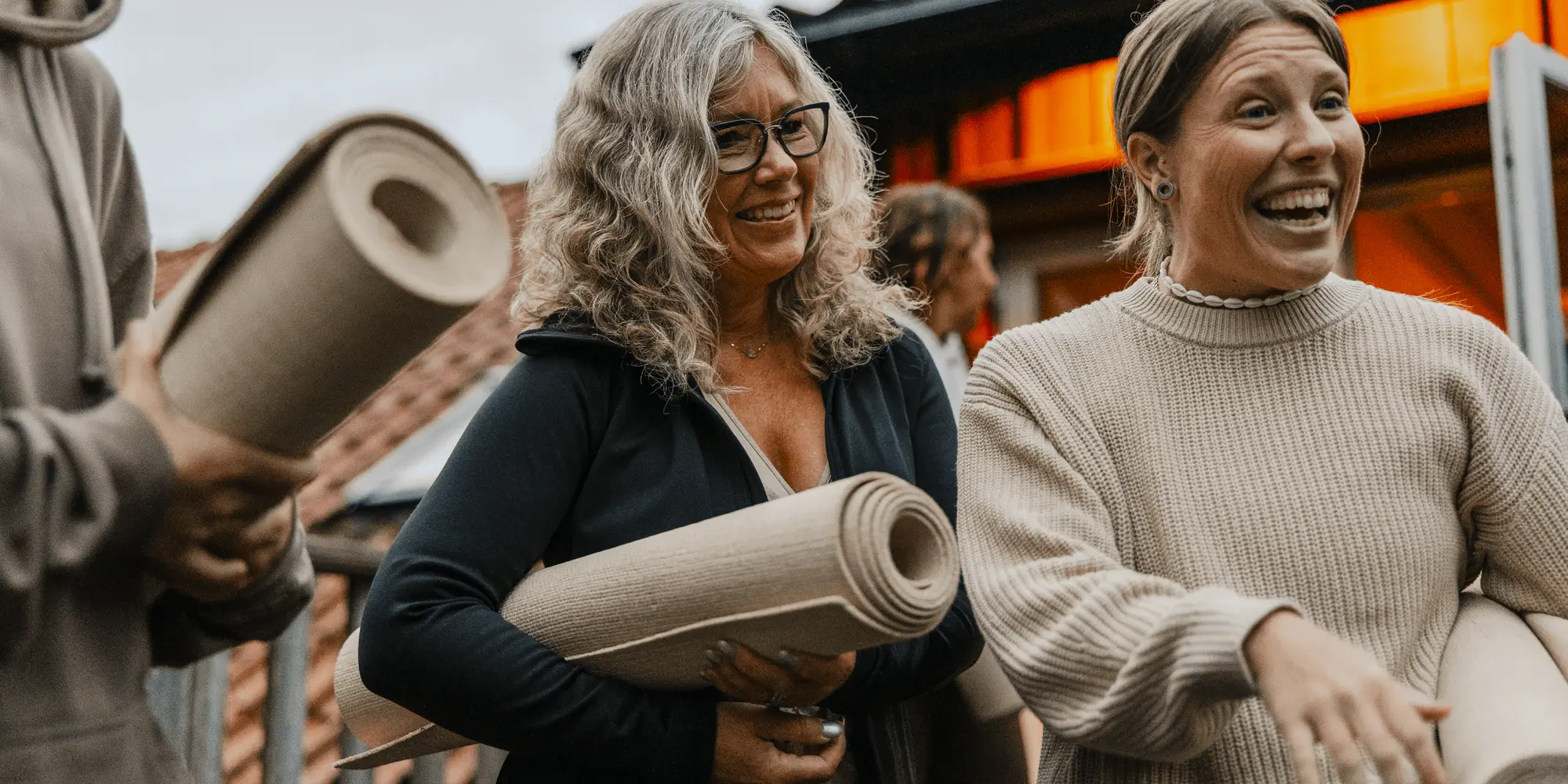 Glada deltagare med yogamattor i händerna samlas utomhus inför en yogaklass. Kvinnorna skrattar och samtalar, omgivna av varm kvällsbelysning och en inbjudande miljö – gemenskap och hälsa i fokus.