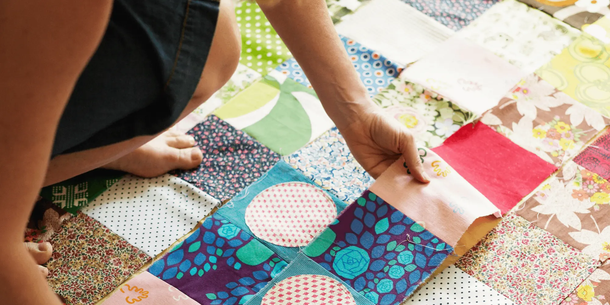 Kvinna sitter på golvet och syr ihop färgglada lappar till ett lapptäcke i ett patchwork-mönster.