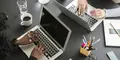 Två personer arbetar vid en svart arbetsyta med varsin laptop, anteckningsbok, kaffekopp och pennor