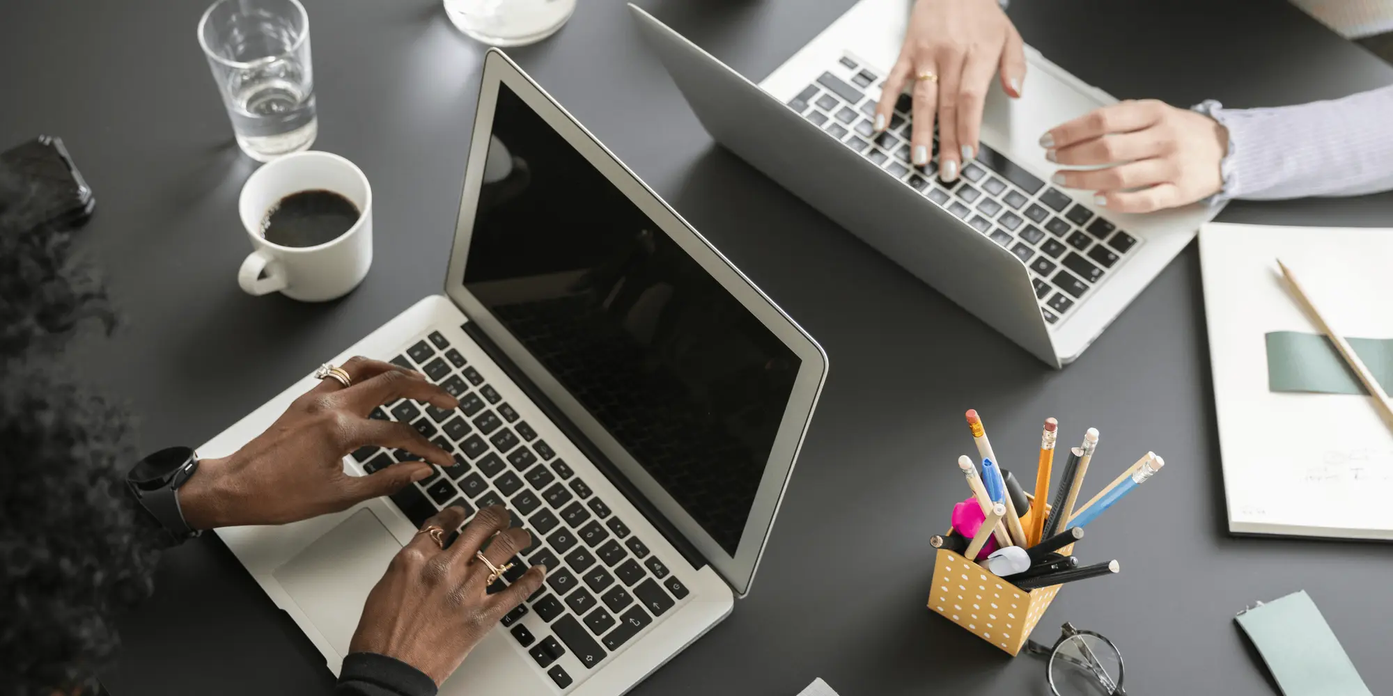 Två personer arbetar vid en svart arbetsyta med varsin laptop, anteckningsbok, kaffekopp och pennor