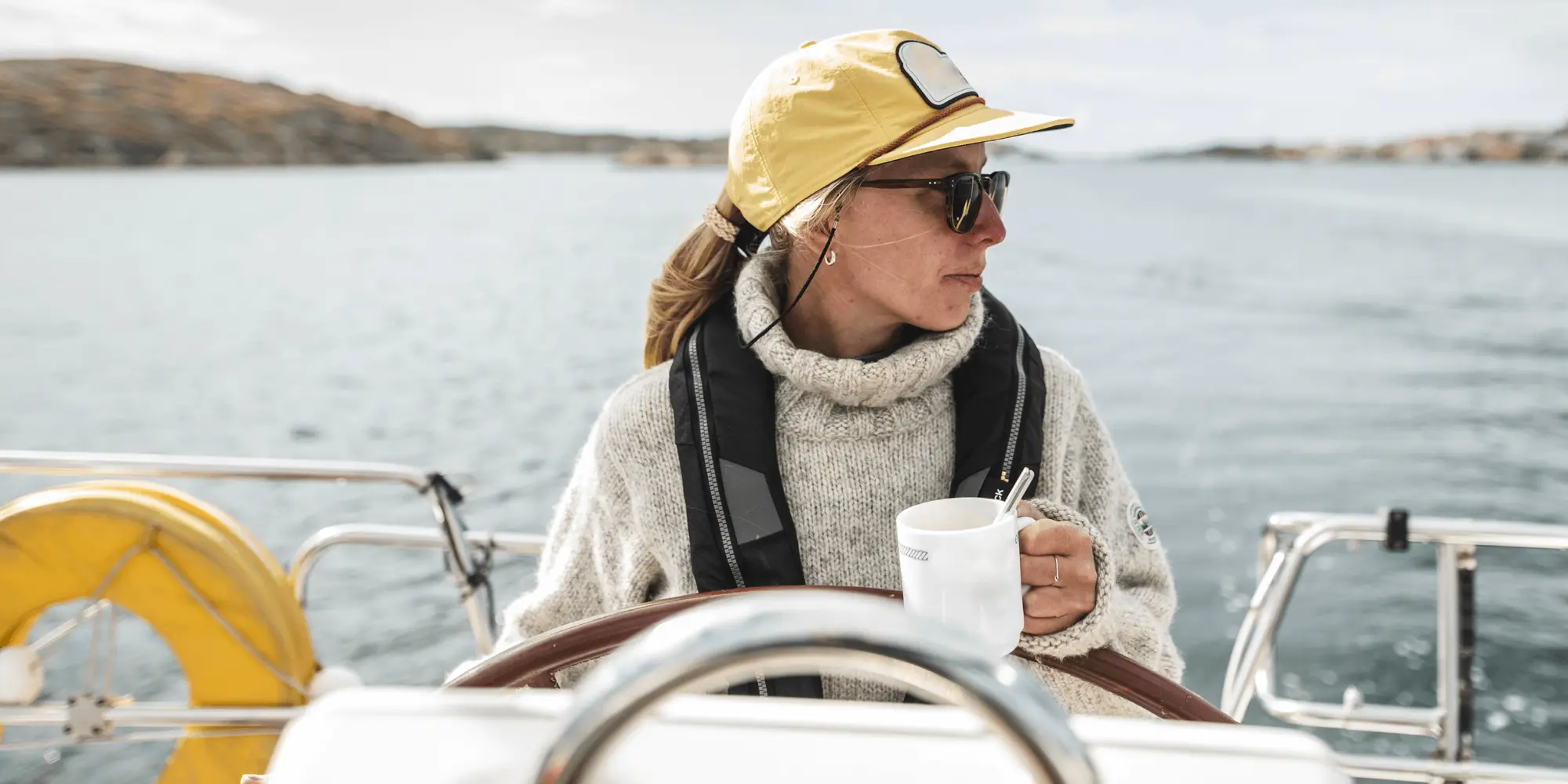 Kvinna i gul keps och stickad tröja styr en segelbåt medan hon dricker kaffe. I bakgrunden syns hav, kobbar och en svensk flagga som vajar i vinden