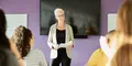 Leende kvinnlig föreläsare med papper i handen står framför en stor skärm och pratar till en grupp elever i ett klassrum