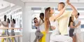Flera par dansar tillsammans i en dansstudio med stora speglar och fönster. Personerna bär träningskläder och dansar i nära position, troligen pardans som salsa eller bachata