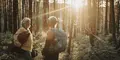 Två kvinnor med ryggsäckar och packning står i en solbelyst skog och samtalar under en vandring