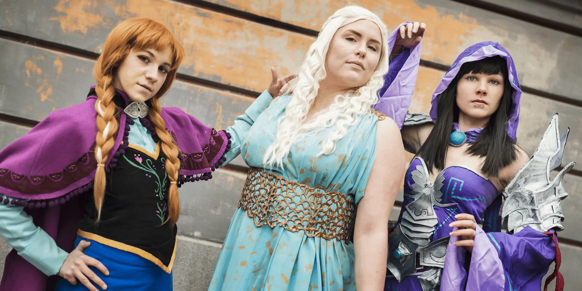 Tre personer i färgstarka och fantasifulla cosplaydräkter poserar framför en vägg – en som Anna från 'Frost', en i blå klänning med långt vitt hår, och en i lila huva och rustningsdetaljer