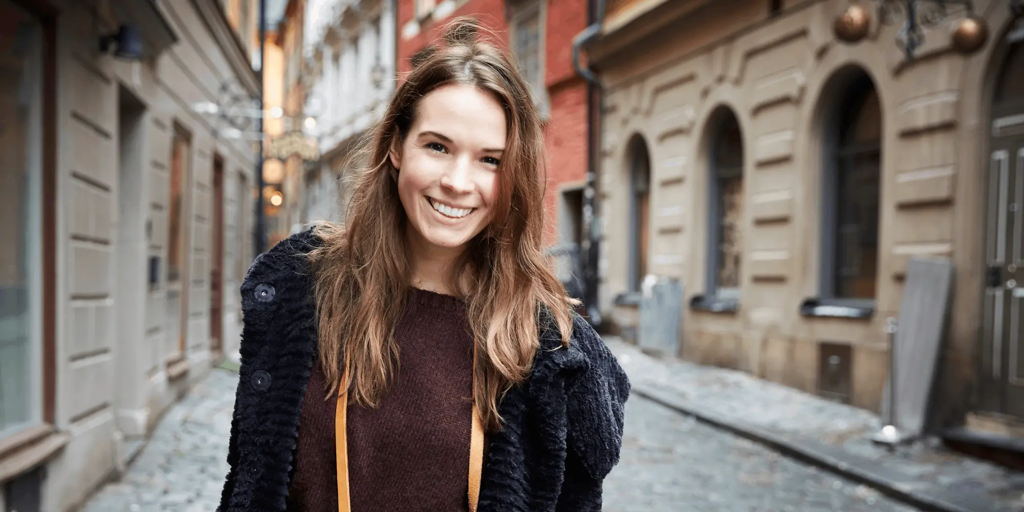 Leende kvinna med kamera runt halsen står på en kullerstensgata i en gammal stadsmiljö