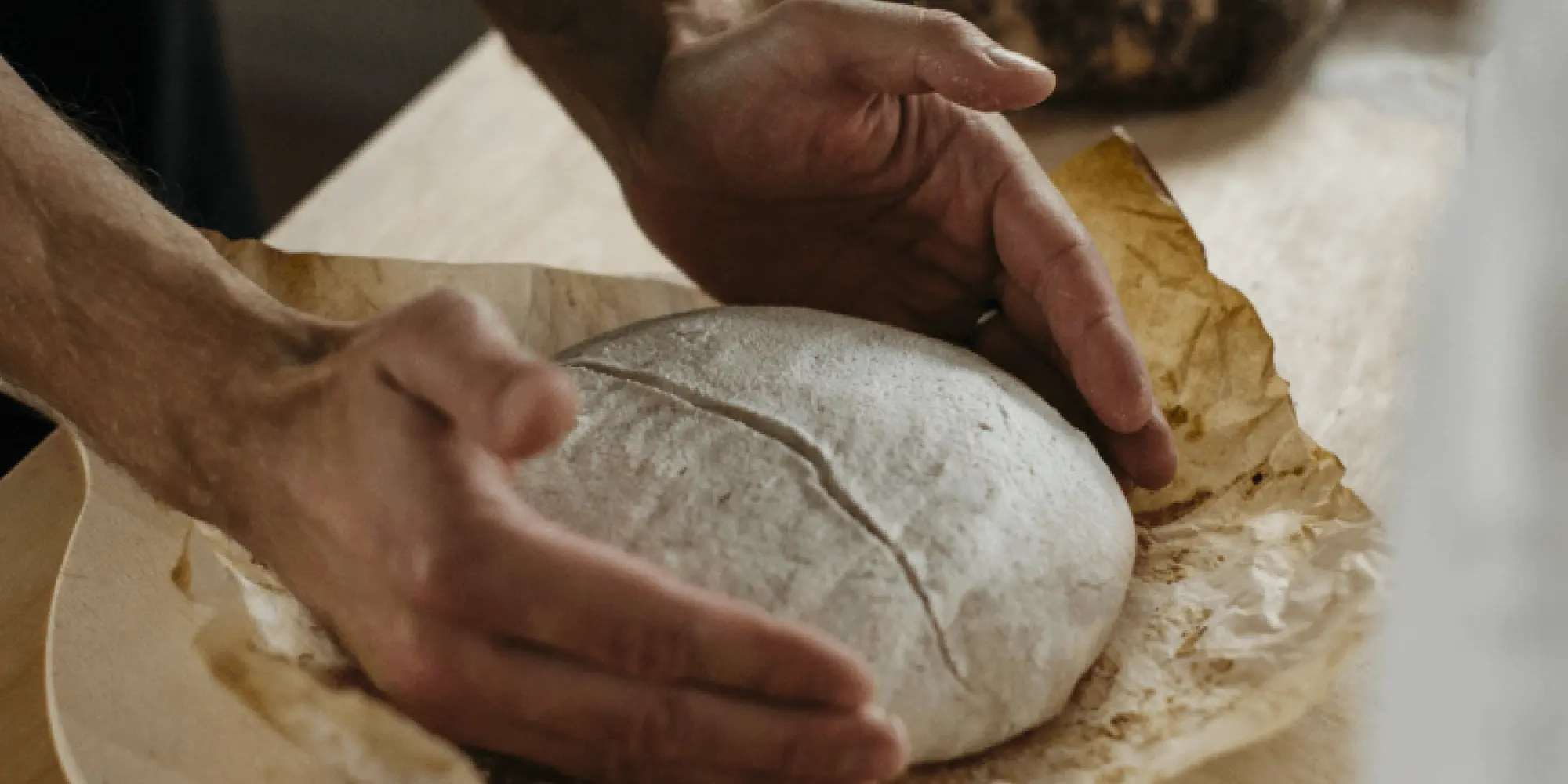 En person formar en deg till ett surdegsbröd på ett bakplåtspapper, redo att gräddas. I bakgrunden syns ett färdigbakat mörkt bröd på en skärbräda.