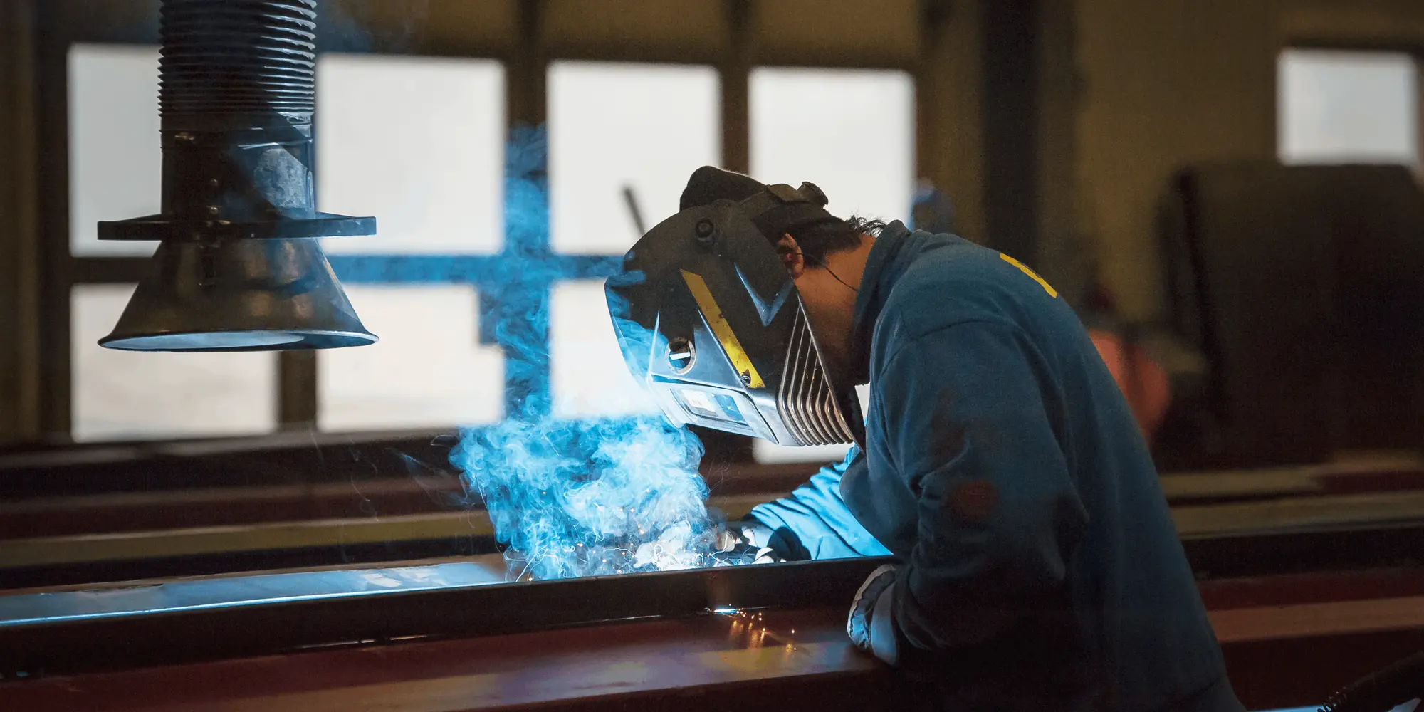 En person iförd svetshjälm och blå arbetskläder svetsar metallbalkar i en verkstad.