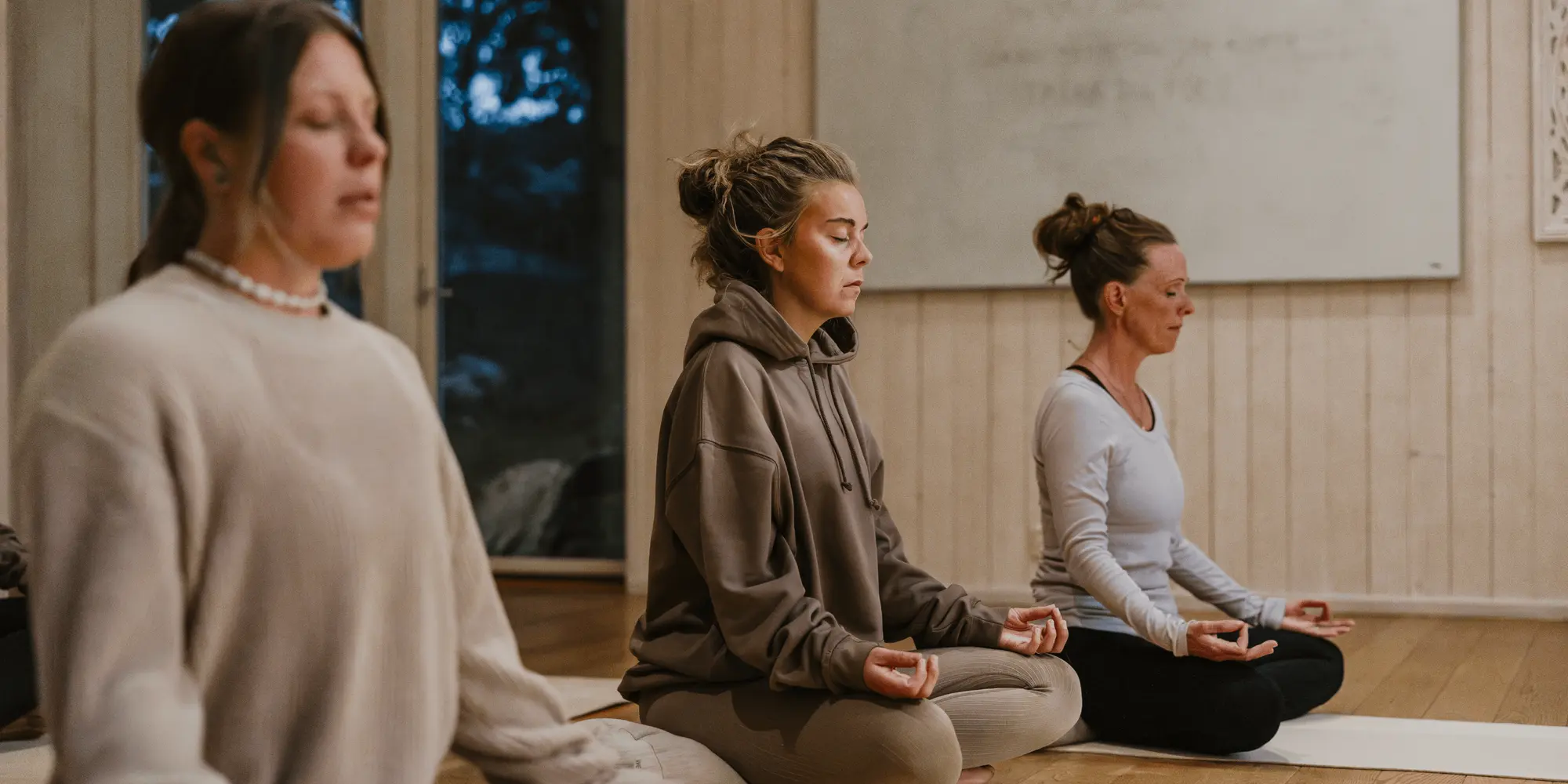 Tre kvinnor sitter i meditationsställning på yogamattor i ett lugnt inomhusrum med trägolv och vita väggar.